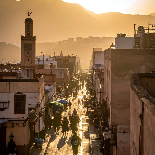 Eine belebte Straße in Tanger, Marokko, getaucht in das warme Licht der untergehenden Sonne, die die einzigartige Architektur und die pulsierende Atmosphäre der Stadt zur Geltung bringt.